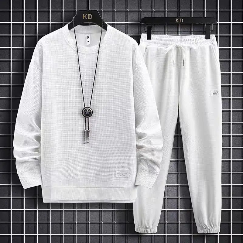 White tracksuit with long-sleeve top and pants on a hanger against a black tiled wall.