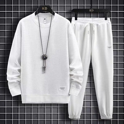 White tracksuit with long-sleeve top and pants on a hanger against a black tiled wall.